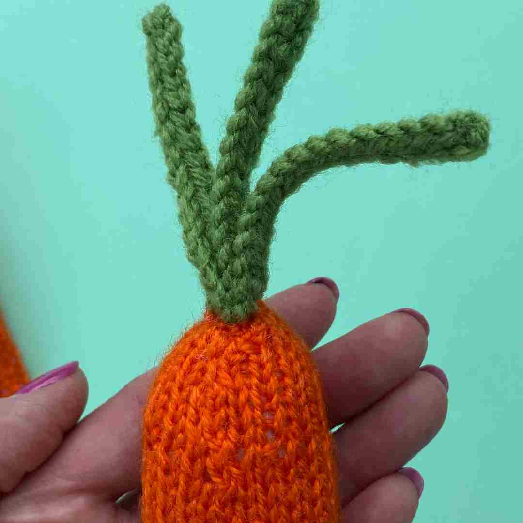 Hand holding top of knitted carrot made from free knitted carrot pattern. Showing the green knitted stalks.