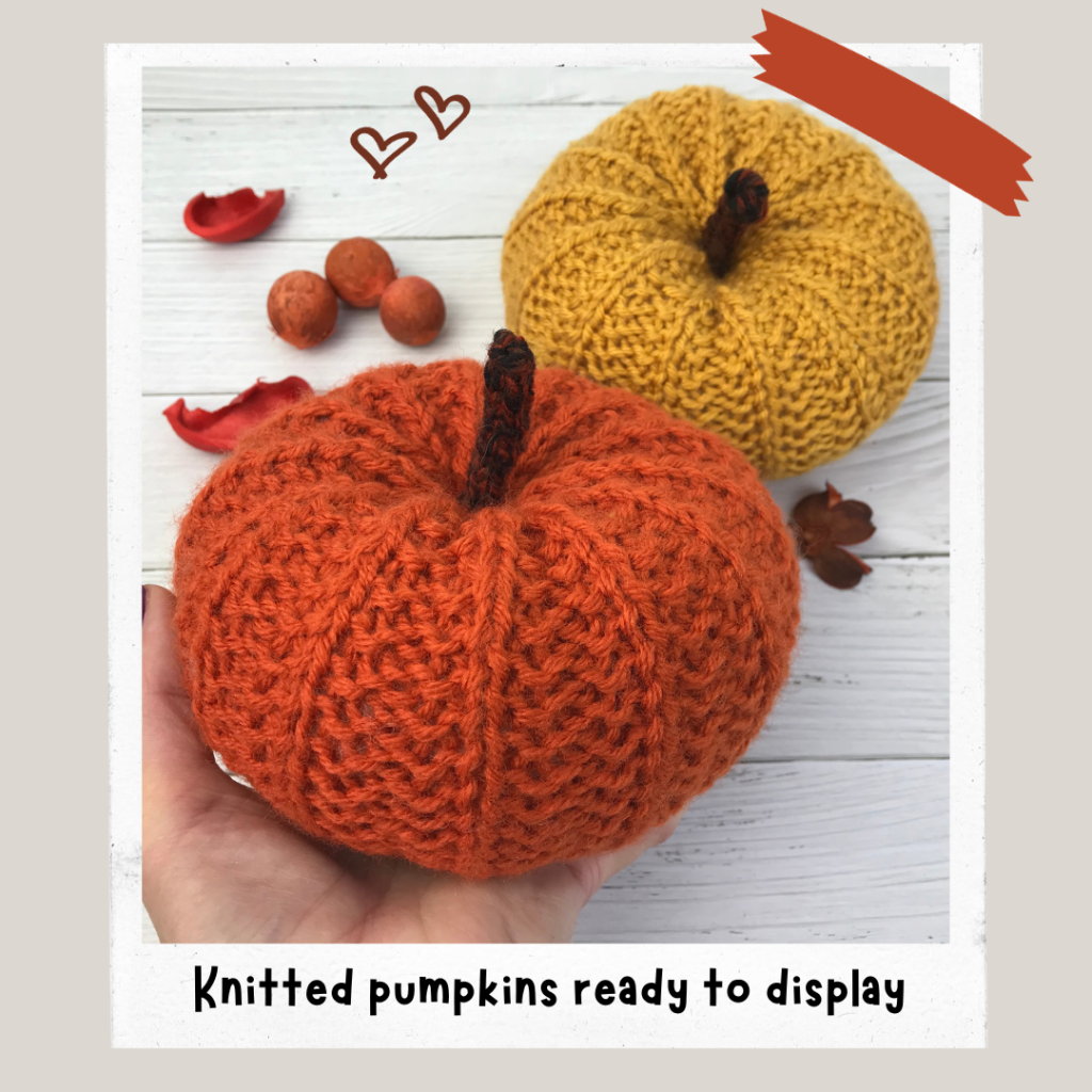 Hand holding a knitted pumpkin in Orange yarn, with a Mustard pumpkin in the backgrounds and acorns scattered on the table