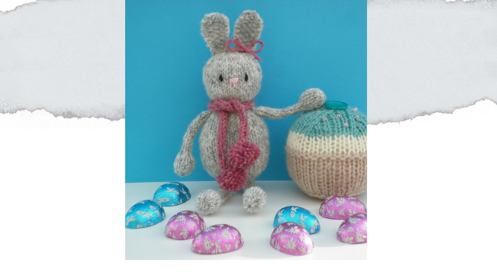 Knitted easter bunny in grey yarn. with a lilac scarf with tiny pompoms. Sat next to a knitted cake with chocolate eggs scattered around.
