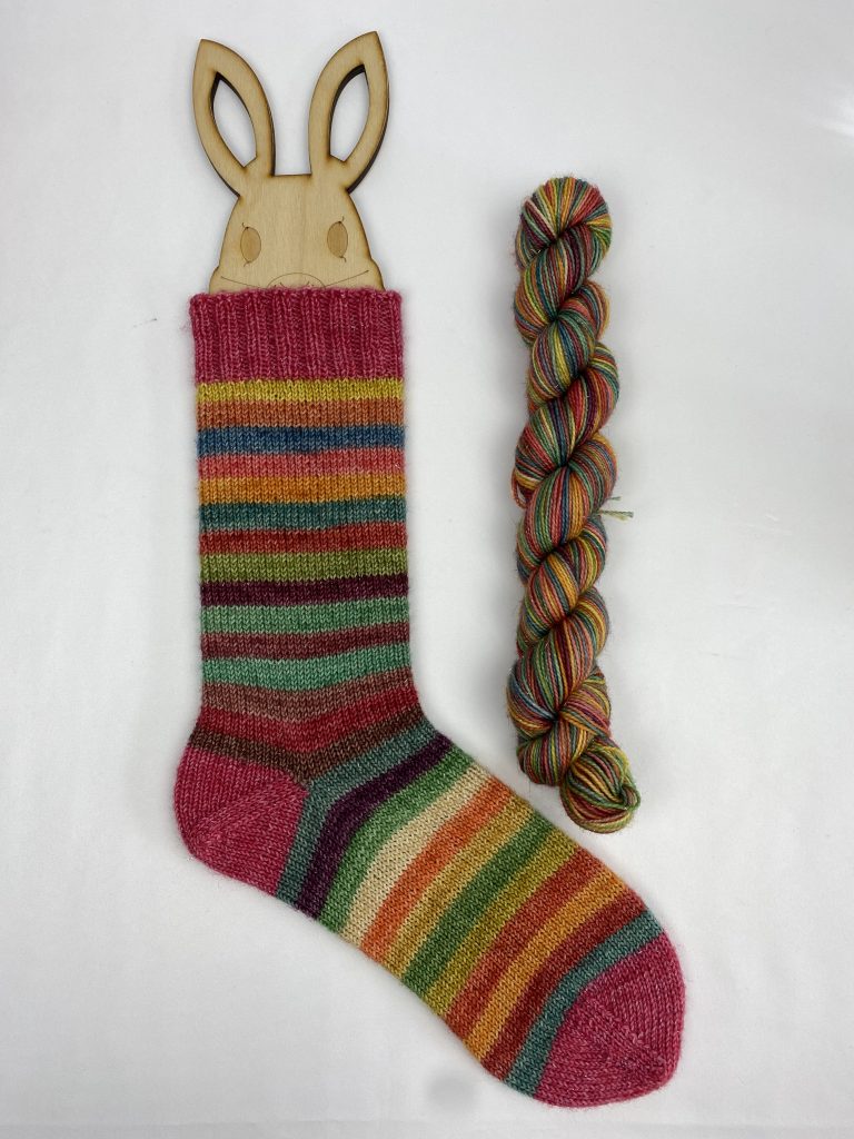 A skein of hand dyed yarn next to a finished sock on a sock blocker, that has been knitted with the yarn, showing the stripes