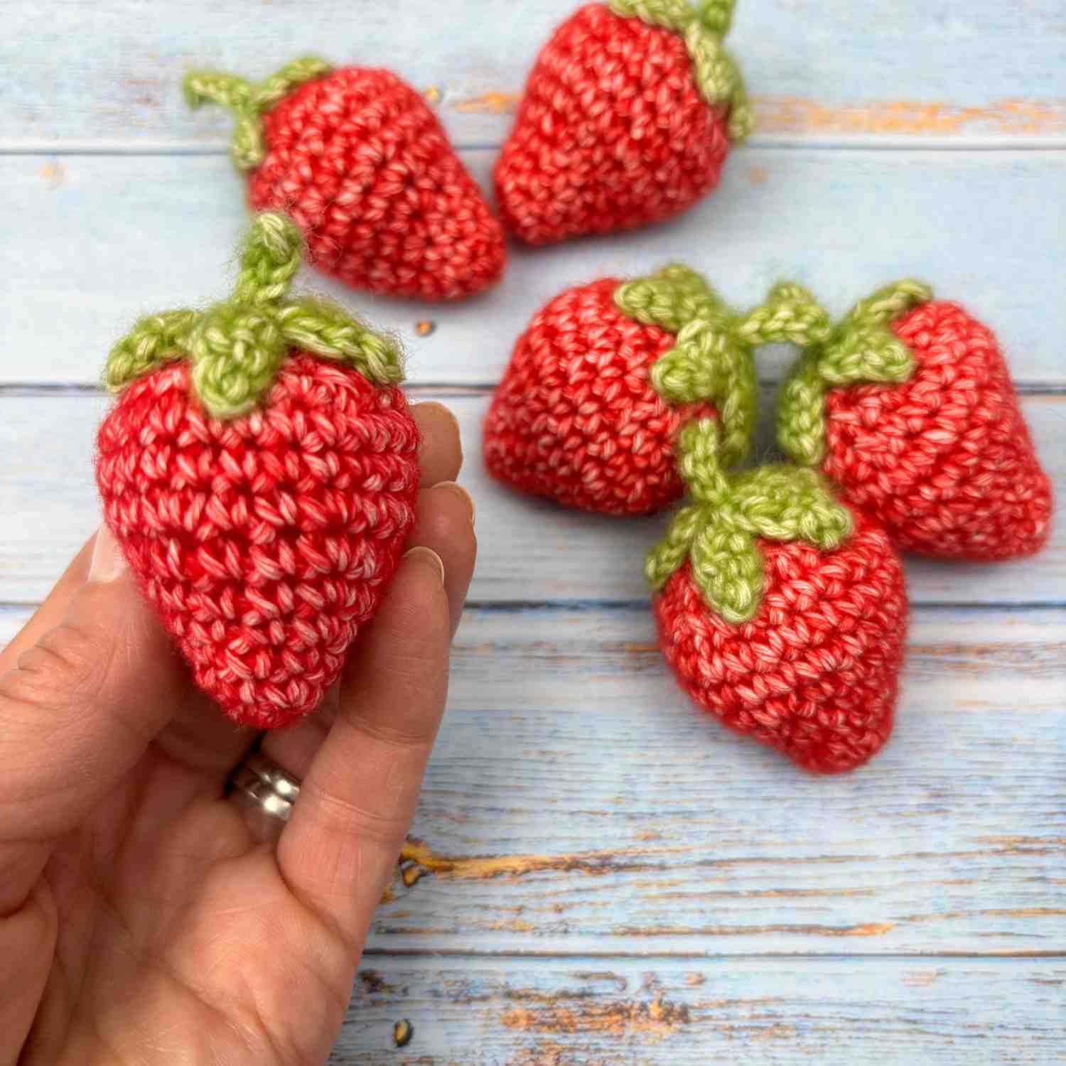 Cute crochet strawberries in red yarn with green leaves, and a hand holding one strawberry. 5 are scattered in the background.