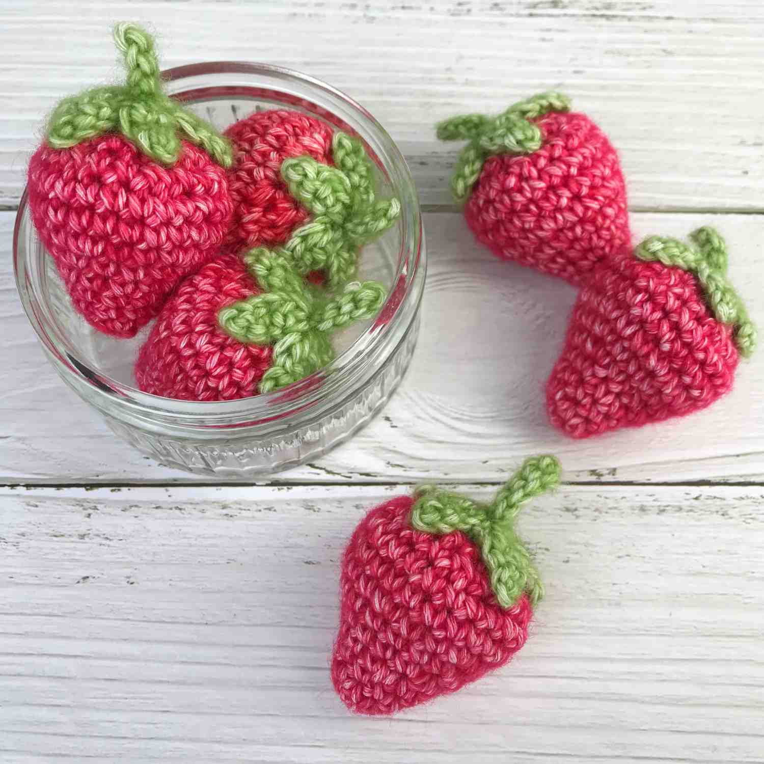 Crochet strawberries in red yarn with green leaves. 3 are in a glass bowl and 3 are scatted around.