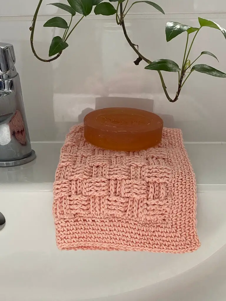 Crochet washcloth on side of sink, folded in a square with a bar of soap on top.