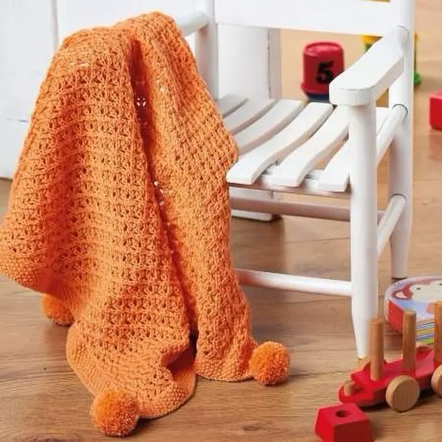 Orange knitting baby blanket made from free baby blanket knitting pattern,  with pompoms, thrown over a white chair.