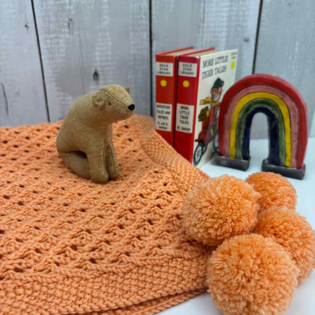 Orange baby blankets made from free baby blanket knitting pattern. Folder to show pompoms, with a little bear sat on top.