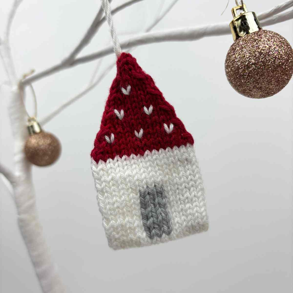 knitted house with a red roof, and grey door, hanging on a white tree.