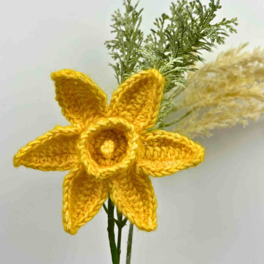 Crochet daffodil flower on a stem with fern leaf at back.
