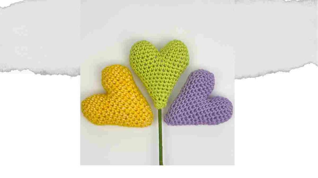 Three amigurumi hearts in yellow, green and lilac. Centre heart is on a stick to put in a plant pot.