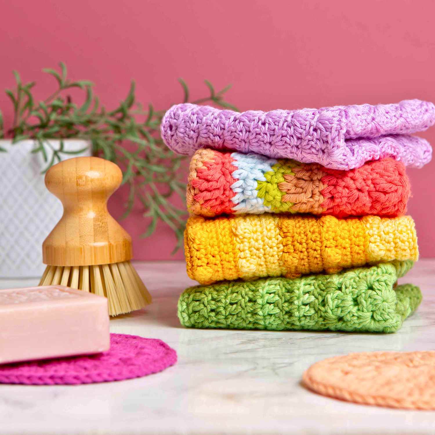 Crochet dishcloths in colourful yarn, stacked on kitchen worktop, with soap and a scrubbing brush. Made from crochet dishcloth patterns Twenty Toi Make series.

