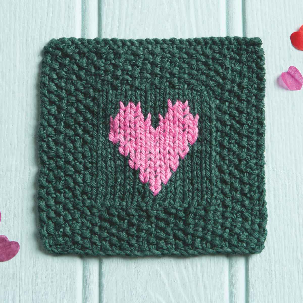 Single knitted square scrubby pattern with heart motif in centre in duplicate stitch, shown in Green cotton yarn with pink heart.