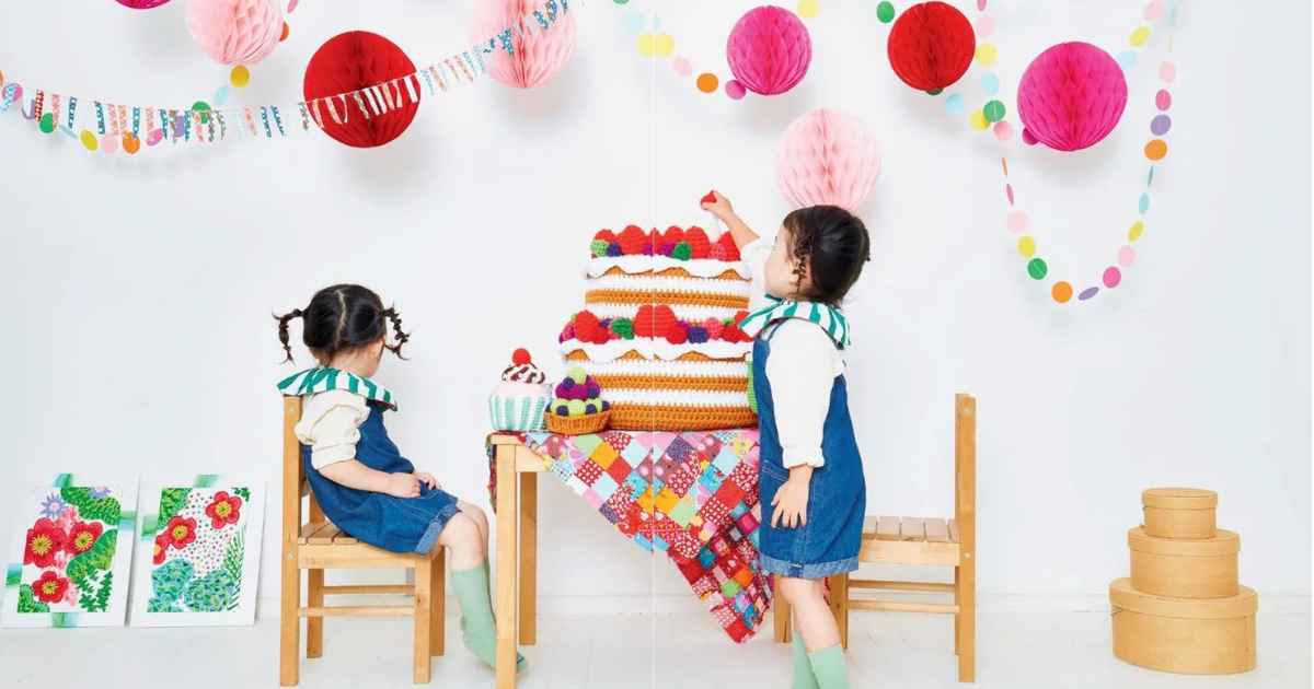 Two small children sat at a table with a huge amigurumi cake and a few smaller crochet cakes.