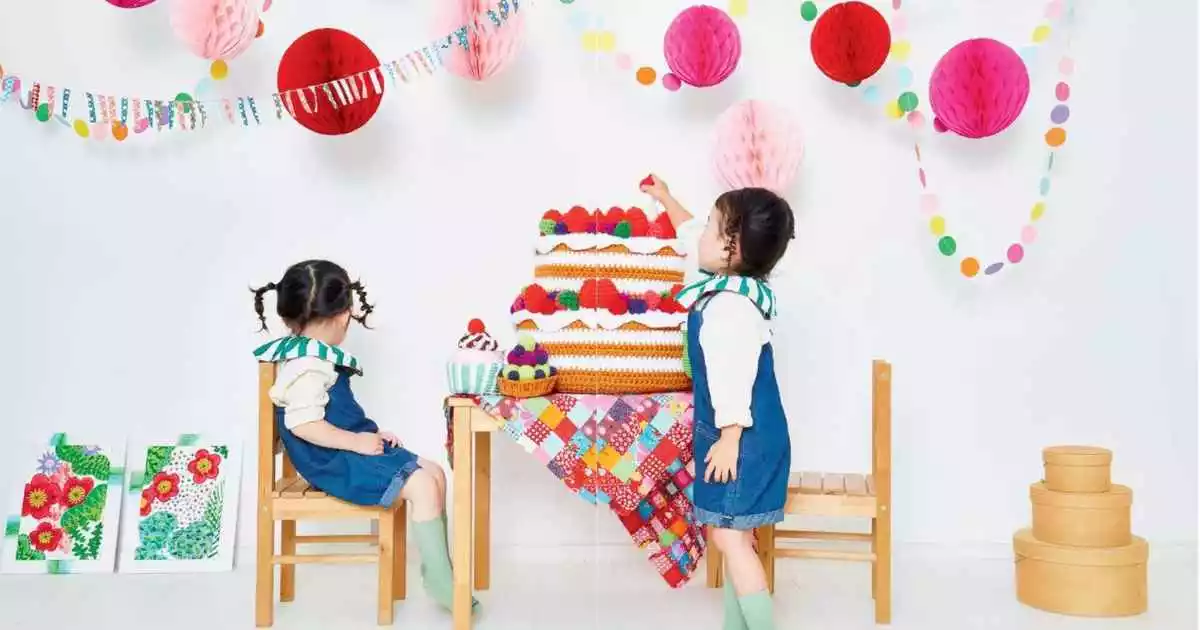 Two small children sat at a table with a huge amigurumi cake and a few smaller crochet cakes.
