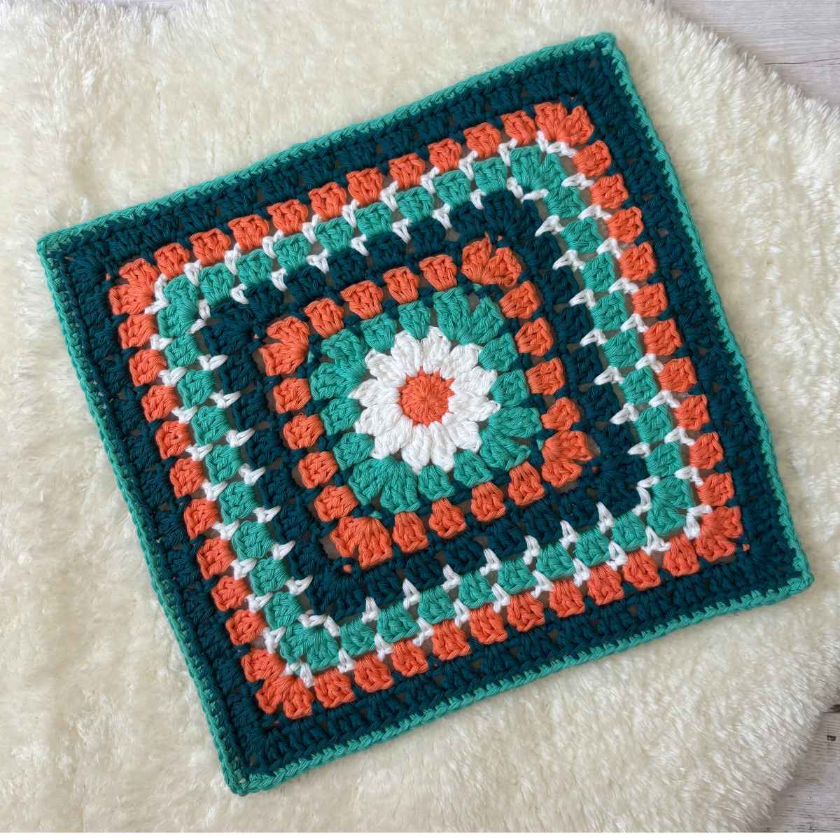 12-inch sized crochet granny square with a daisy granny centre surrounded by a framed Block stitch pattern. In Duck Egg Blue yarn with Teal, White and Coral.